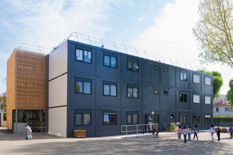  Bâtiment de groupe scolaire modulaire moderne ALGECO® à trois étages avec façade métallique foncée, côtés blancs et habillage en bois pour l’escalier gauche